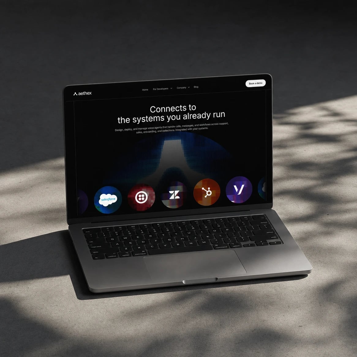 aethex website: Laptop on grey concrete in leaf shadow, showing the Aethex integrations section 'Connects to the systems you already run' with round integration logos