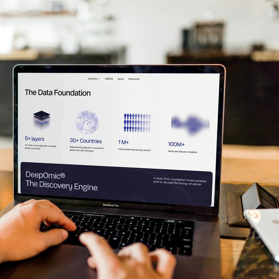 Nexomic website: Laptop on a café table showing Nexomic's 'The Data Foundation' section and the DeepOmic engine