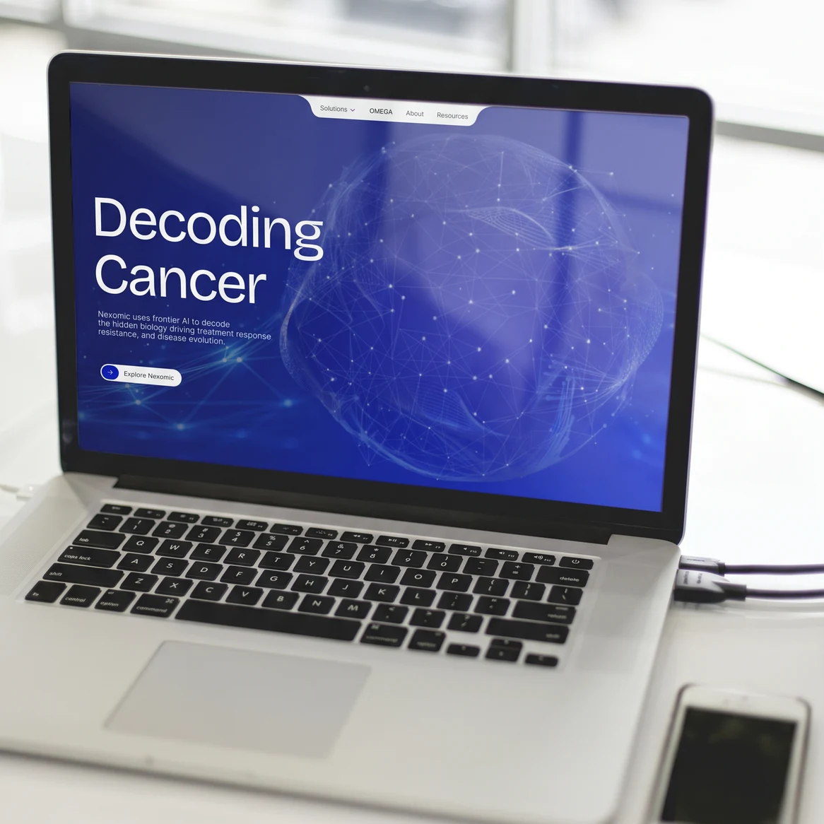 Nexomic website: Laptop on a bright desk by a window, showing the Nexomic 'Decoding Cancer' hero